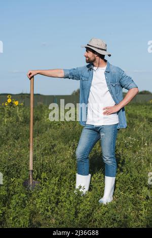 vista completa dell'agricoltore felice con pala in piedi con mano sull'anca in campo Foto Stock