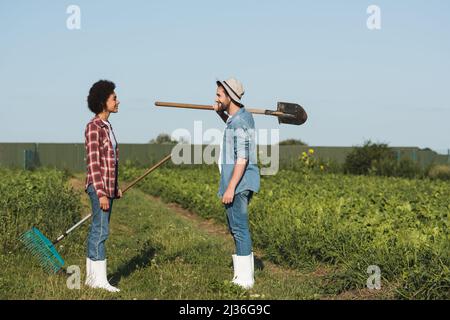 vista laterale di felici agricoltori interrazziali con pala e rastrelli che si guardano a vicenda su terreni agricoli Foto Stock