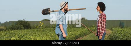 allegri agricoltori multietnici con strumenti di giardinaggio che si guardano a vicenda nel campo, banner Foto Stock