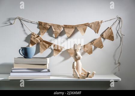 Pila di libri, penne in un portamatite, penne in feltro, figurine in legno di una lepre sono su scaffale. Garland di bandiere di carta del mestiere sul muro sopra la lei Foto Stock