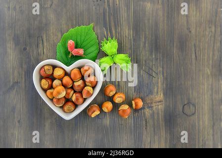 Nocciole fresche non sgusciate in un piatto a forma di cuore con foglie verdi in un piano in alto stendere la vita su un tavolo di legno con copyspace Foto Stock