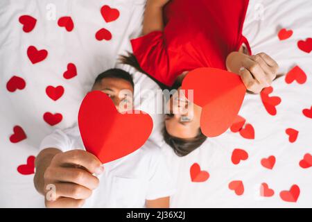 Vista dall'alto della giovane coppia attrattiva in amore che si trova sul letto e mostra cuori di carta rossa. Buon San Valentino concetto di giorno Foto Stock