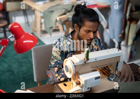 Scatto ad angolo alto di giovane designer di etnia indiana seduto al tavolo utilizzando la macchina da cucire per fare l'abito per la nuova collezione Foto Stock