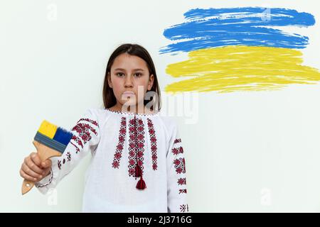 Una ragazza in un abito ricamato si erge contro una parete bianca, tenendo una spazzola dipinta di blu e giallo. La ragazza protesta contro la guerra. Ucraino-Russi Foto Stock