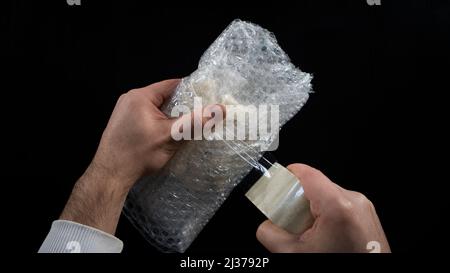 Imballaggio con nastro per condotti e bolle, confezione per spedizione, imballaggio per mani maschili, isolato su sfondo nero, vista dall'alto Foto Stock