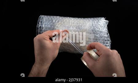 Imballaggio con nastro per condotti e bolle, confezione per spedizione, imballaggio per mani maschili, isolato su sfondo nero, vista dall'alto Foto Stock