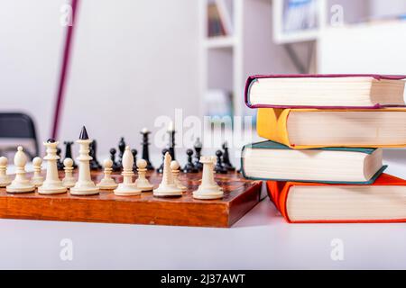 Sul tavolo bianco si trova una vecchia scacchiera di legno con pezzi di scacchi bianchi e neri disposti. Nelle vicinanze si trova una pila di libri multicolore. Tutto è letto Foto Stock