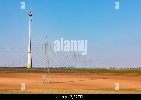 Diversi tralicci di elettricità dominano il paesaggio rurale con un'enorme torre a turbina eolica sullo sfondo Foto Stock