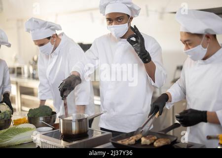 Il team multirazziale dello chef cuoca in uniforme bianca cucinando insieme in cucina. Ragazzi asiatici, latini ed europei che cucinano insieme. Cuochi che indossano maschere per il viso e guanti protettivi Foto Stock