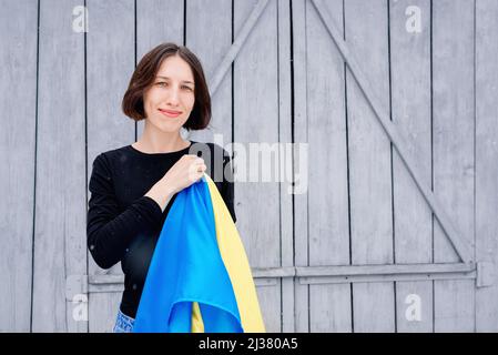 Giovane ragazza che tiene una bandiera di stato blu e gialla dell'Ucraina nella sua mano destra sopra un cuore su uno sfondo di recinto grigio di legno. Una donna in un jac nero Foto Stock