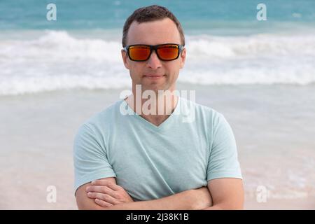 Ritratto di giovane adulto sportivo sorridente uomo europeo in occhiali da sole rossi lucenti, foto all'aperto scattata su una spiaggia estiva Foto Stock