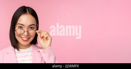 Ritratto di donna d'affari asiatica in occhiali, guardando intrigato alla macchina fotografica e sorridente, professionista donna di vendita fissando con interesse, sfondo rosa Foto Stock