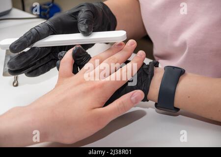 Mani gloved di un manicurist esperto che archivano le unghie di una donna giovane con una lima del chiodo. Mani durante una sessione di cura manicure in un salone di spa. Manicurista fi Foto Stock