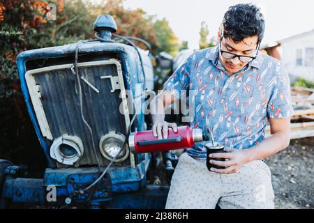 Giovane latino che serve compagno con un thermo rosso, in uno spazio naturale, accanto ad un vecchio piccolo trattore blu. Bevanda latina. Foto Stock