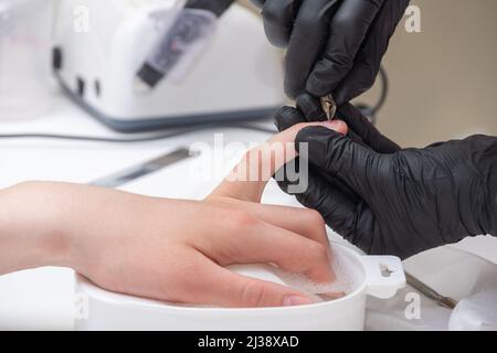 Manicurista che rimuove la cuticola dalle unghie femminili da un pestello di metallo durante l'immersione delle unghie nel bagno al salone delle unghie. Mani durante una cura manicure sessi Foto Stock