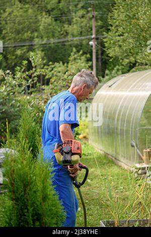 L'operatore con un tagliabasette in mano rasa l'erba davanti alla casa. Il rifinitore è nelle mani di un uomo. Il giardiniere taglia l'erba. Sollevi Foto Stock