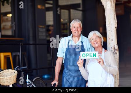 Vieni a vedere cosa abbiamo da offrire. Ritratto di una coppia anziana felice che tiene un segno aperto nel loro negozio. Foto Stock