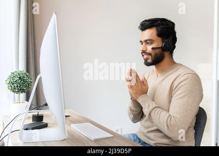 Uomo indiano misto da corsa che indossa cuffie wireless in elegante camicia casual utilizzando un computer portatile mentre si siede alla scrivania. Ha ispirato un dipendente indù maschio in cuffie con microfono facendo gesto di saluto namaste Foto Stock