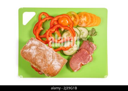 Pomodori a fette, peperoni, cetrioli, prosciutto e pane su un tagliere verde. Isolato su bianco, tracciato di ritaglio incluso Foto Stock