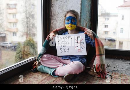 Una bambina con una bandiera Ucraina dipinta sul suo volto è seduta sotto una coperta con un poster cielo chiuso. Bambini e guerra. Foto Stock