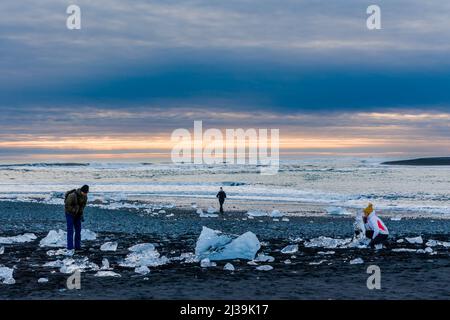 JOKULSARLON,ISLANDA - NOVEMBRE 18 2021: Turisti che esaminano iceberg e cristalli sulla spiaggia di sabbia nera a Jokulsarlon, Islanda Foto Stock