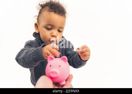 Adorabile ragazzo biraciale focalizzato bambino mettendo le sue monete nella banca rosa piccante su sfondo bianco. Foto di alta qualità Foto Stock