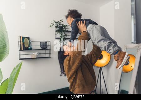 Giovane mamma caucasica dai capelli scuri che solleva il suo adorabile figlio biraciale nel loro appartamento moderno. Relazione tra padre e figlio. Foto di alta qualità Foto Stock