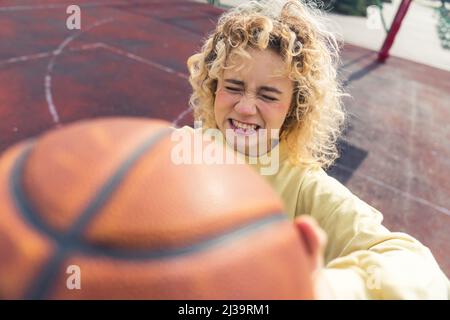 Giovane donna ippica bionda che mostra una palla di basket alla macchina fotografica, facendo il volto sciocco, in piedi sul campo sportivo primo piano copia spazio . Foto di alta qualità Foto Stock