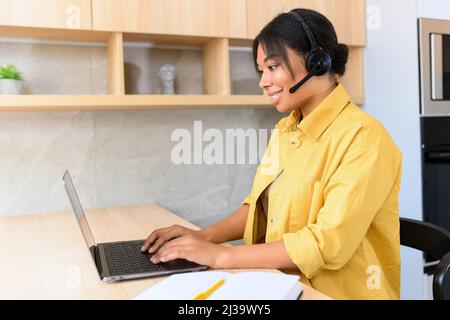 Sorridente giovane donna multirazziale attraente usa la cuffia per parlare in linea lavorando a distanza in ufficio domestico. Vista laterale di una donna etnica che utilizza cuffie con microfono per la comunicazione online Foto Stock