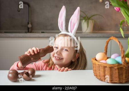 Carino bambina indossa orecchie di coniglietto mangiando cioccolato il Coniglio di Pasqua. Kid giocando caccia all'uovo di Pasqua. Adorabile bambino celebrare la Pasqua in casa. Foto Stock