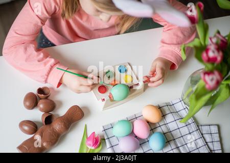 Vista dall'alto di una bambina con orecchie conigliate che tengono il pennello e decorano le uova di pasqua con acquerelli. Tulipani, cioccolato pasquale e uova su un bianco Foto Stock