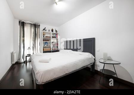 camera da letto con libreria bianca, finestra con tende grigie e letto matrimoniale con vestiti bianchi e asciugamani puliti Foto Stock
