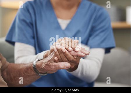 Anche gli eroi indossano scrub. Shot di un'infermiera irriconoscibile che tiene una mano del paziente durante un controllo a casa. Foto Stock