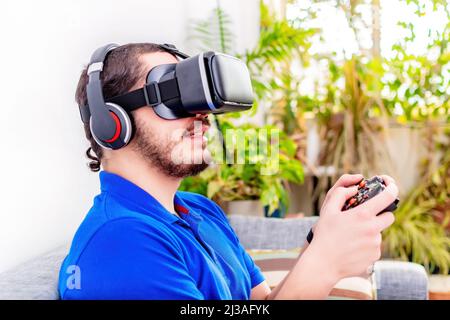 Un giovane bearded medio-orientale che usa la cuffia per realtà virtuale, il controller e gli occhiali a casa Foto Stock