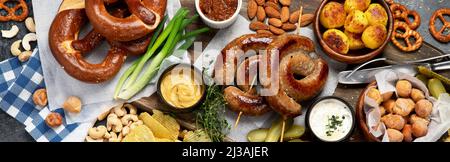 Cena all'Oktoberfest con salsicce di maiale alla griglia, pasta pretzel, patate, insalata, spuntini, salse, birra su sfondo scuro, vista dall'alto. Foto Stock