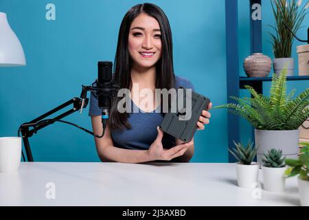 Creatore di contenuti asiatici con v mount power batteria di registrazione podcast per il canale di vlogging presentazione di apparecchiature di produzione videografica. Social media influencer revisionando adattatore portatile Foto Stock