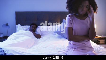 Non riesco a dormire con questo mal di testa fremolante. Scatto di una giovane donna interessata che si siede su un letto con il marito che dorme in Foto Stock