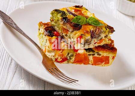 una porzione di frittata di verdure al forno con uova sbattute, cipolla rossa, patate dolci, broccoli, pepe rosso e funghi su piatto bianco su legno bianco Foto Stock