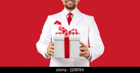 Giovane uomo in un abito in piedi in studio e tenendo una scatola regalo bianco legato con un arco rosso Foto Stock