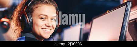 Vista laterale di una ragazza felice gara mista, giocatore di cybersport femminile che indossa le cuffie sorridendo alla fotocamera durante la partecipazione al torneo di eSport Foto Stock