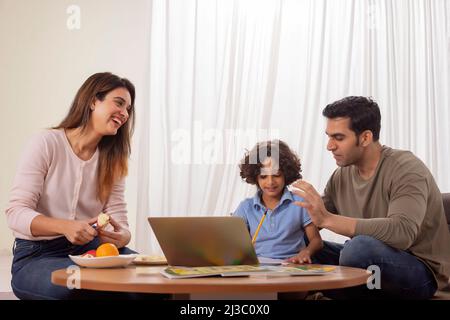 Padre che aiuta il figlio con i suoi compiti e la madre che taglia i frutti accanto Foto Stock