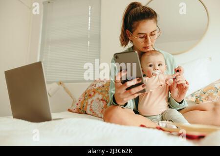 Equilibrare maternità e lavoro. Nuova mamma che prende una penna dal suo bambino mentre lavora su un letto. Single madre multitasking cercando di leggere un messaggio di testo Whi Foto Stock