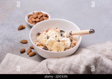 Farinata d'avena con fette fresche di banana, mandorle, nocciole e gocce di cioccolato per una colazione sana su sfondo grigio di cemento Foto Stock