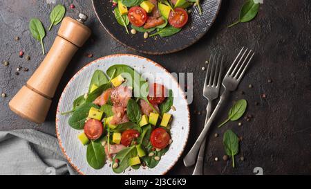 Vista dall'alto di cibi sani. Insalata con foglie di spinaci freschi, pomodori ciliegini, avocado a fette e pesce salato di salmone rosso su fondo di cemento scuro Foto Stock