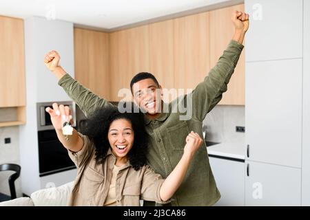 Giovane coppia multirazziale sposata sorridendo allegro e mostrando le chiavi di un nuovo appartamento, abbracciando e guardando la macchina fotografica, mentre si trova in posizione moderna cucina di nuova casa. Concetto immobiliare e familiare Foto Stock