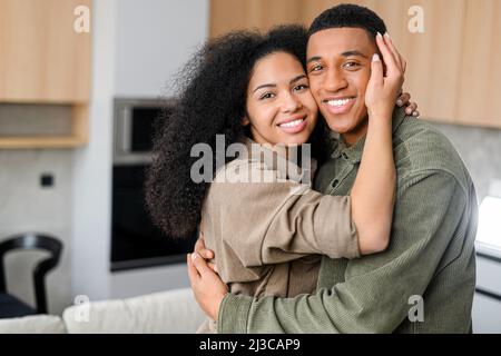 Bella coppia multirazziale in abbraccia in accogliente cucina moderna a casa. Sorridente ragazzo è abbracciante ragazza affascinante. Concetto di amore e affetto Foto Stock