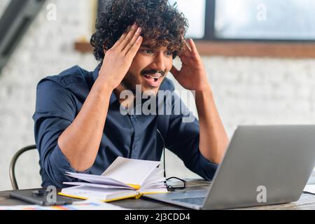 Stanco dipendente indiano maschile ufficio urlando mentre si guarda lo schermo del laptop, sensazione di noioso e arrabbiato. Annoiato uomo orientale in elegante camicia casual seduto alla scrivania Foto Stock