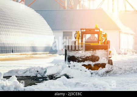 Il trattore sgomma il territorio della base di produzione dalla neve in una giornata invernale limpida. Rimozione della neve dopo la nevicata. Sfondo. Foto Stock