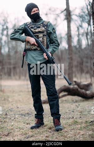 Il giovane uomo si alza con la pistola a pompa nelle sue mani, temuta in corazza e balaclava nella foresta. Foto Stock