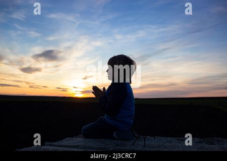 Silhouette di un ragazzo seduto sulle ginocchia, le mani piegate in preghiera, voltandosi verso Dio al tramonto. Bambini contro la guerra, il bambino chiede la pace per il suo hom Foto Stock